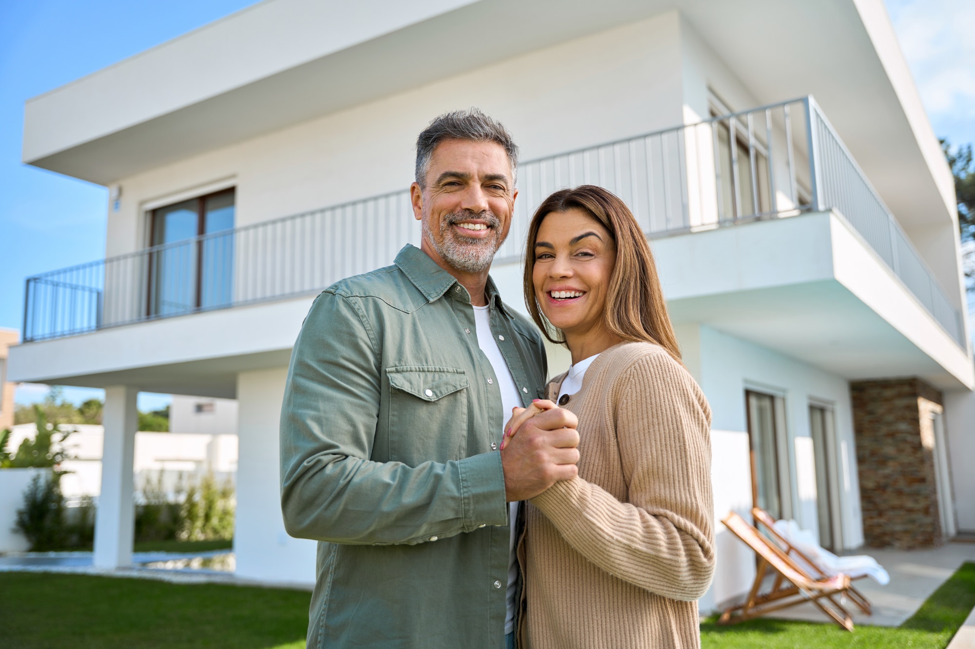Portrait of happy mature family couple property owners standing outside house.